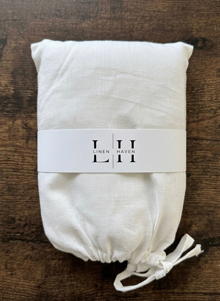 An off-white linen fabric item, likely a pillowcase or folded textile, wrapped in a white paper band printed with the brand name 'LINEN HAVEN' and tied with a drawstring, placed on a dark wood background.